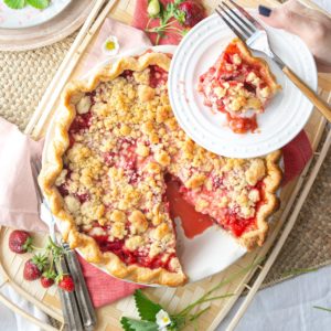 A strawberry rhubarb pie with a slice cut out on a basket and strawberry leaves and flowers.