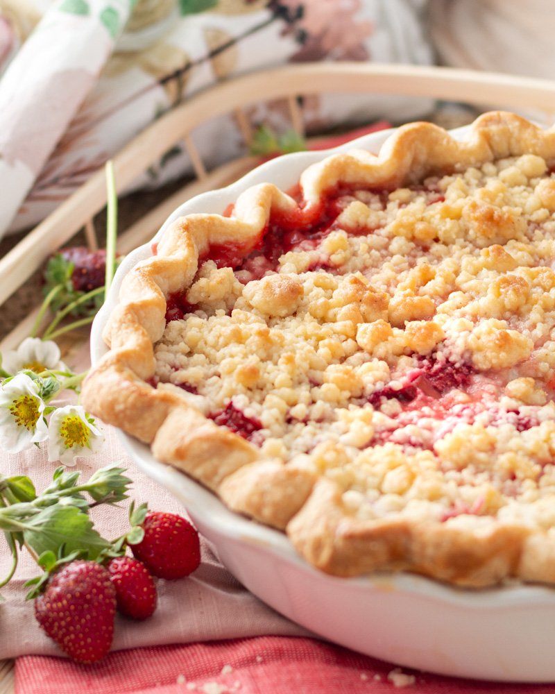 A strawberry rhubarb pie with a slice cut out on a basket and strawberry leaves and flowers.