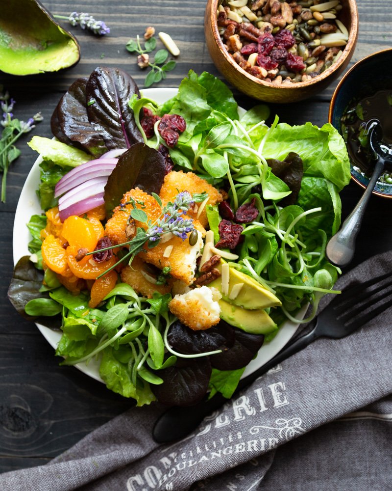 A bowl of crisp salad greens topped with sunflower sprouts, mandarin oranges, crispy fried feta cheese, herby honey, dried cranberries, avocado and red onion.