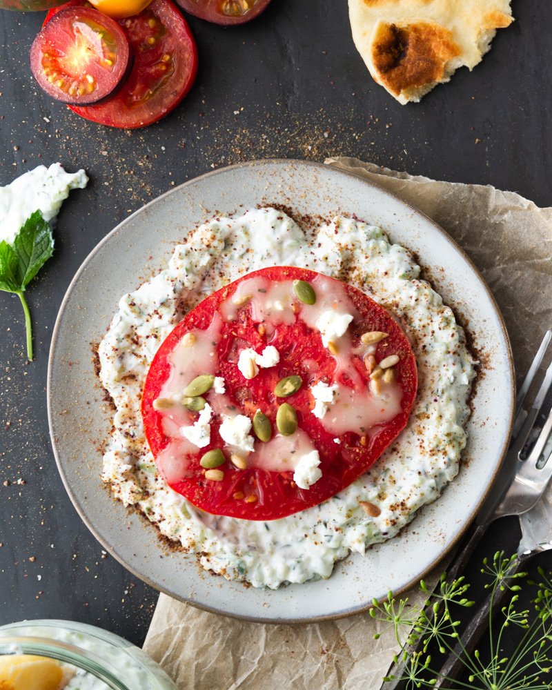 A plate of tzatziki sauce with tomato, pita bread and fresh herbs.