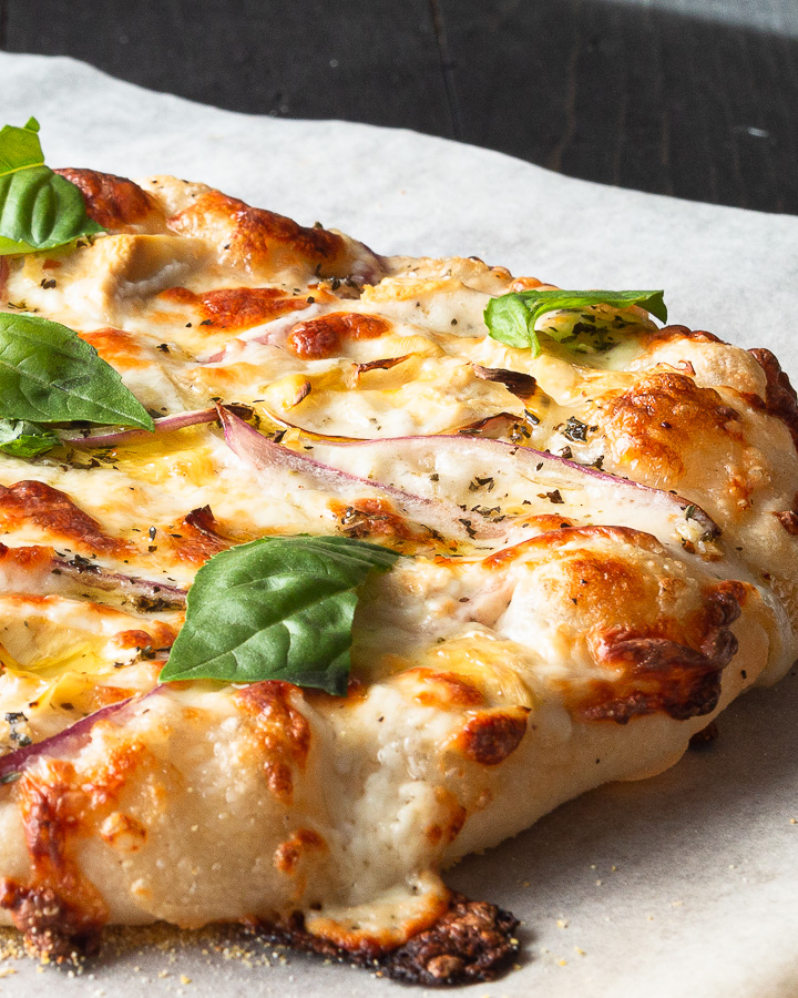 A white pizza using a master pizza dough recipe, Alfredo sauce, red onion, artichoke hearts, fresh basil leaves, and an herb oil to finish.